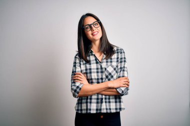 Mavi gözlü, beyaz arka planda gözlüklü, kollarını kavuşturup kameraya gülümseyen esmer bir kadın. Pozitif kişi.