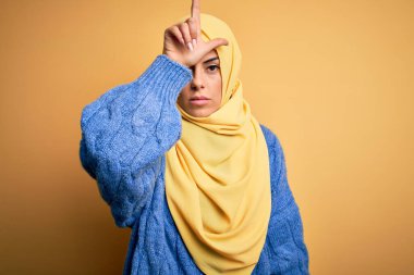 Genç, güzel, esmer, Müslüman kadın izole edilmiş sarı arka plan üzerine Arap başörtüsü takıyor. Alnında parmakları olan insanlarla alay ediyor, alay ediyor, alay ediyor..