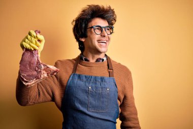 Genç yakışıklı kasap biftekle karşılaşıyor. Sarı arka planda duruyor. Yüzünde gülümseme, doğal bir ifade var. Kendinden emin gülen.