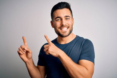 İzole edilmiş beyaz arka plan üzerinde duran sıradan tişört giyen genç yakışıklı adam gülümsüyor ve kameraya bakıyor iki eliyle ve parmaklarıyla kenarı işaret ediyor..
