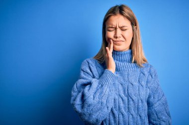 Sarışın, güzel bir kadın mavi yünlü bir arka plan üzerine kazak giyip diş ağrısı ya da diş hastalığından dolayı acı dolu bir ifadeyle ağıza dokunuyor. Dişçi