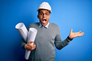 Genç Afrikalı Amerikalı mimar kaskını takmış elinde planlarla çok mutlu ve heyecanlı, kazanan ifade zaferi büyük bir gülümsemeyle kutluyor ve ellerini kaldırıyor.