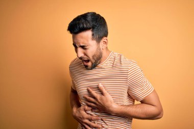 Sakallı genç yakışıklı bir adam sarı arka planda normal çizgili tişört giyiyor. Midesi bulanıyor, acı verici bir hastalığı var. Ache kavramı.