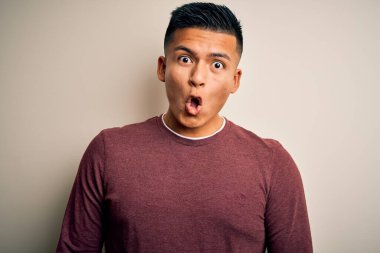 Genç, yakışıklı, latin bir adam, günlük süveter giyiyor, beyaz arka planda dikiliyor, korkmuş ve şok olmuş bir halde..