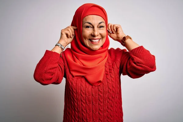 Middle age brunette woman wearing muslim traditional hijab over isolated white background Smiling pulling ears with fingers, funny gesture. Audition problem