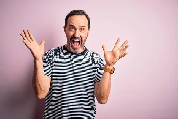 Orta yaşlı, beyaz bir adam, günlük çizgili tişört giymiş, pembe arka plan üzerinde durmuş çılgın ve başarısını kutluyor, kollarını kaldırmış ve gözleri açık bir şekilde heyecanla haykırıyor. Kazanan konsept