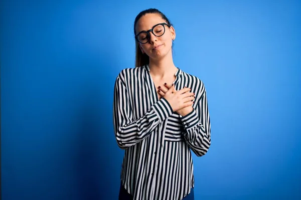 Beautiful blonde woman with blue eyes wearing striped shirt and glasses over blue background smiling with hands on chest with closed eyes and grateful gesture on face. Health concept.
