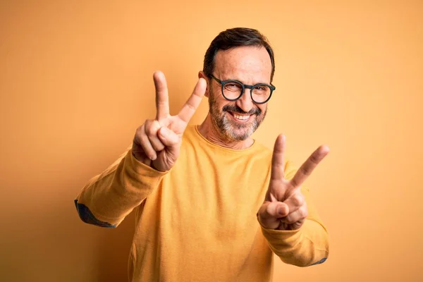 両方の手の指が勝利の印を行うことを示す舌で微笑む孤立した黄色の背景の上にカジュアルなセーターと眼鏡を身に着けている中年の蜂蜜の男 第二番 — ストック写真