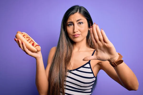 若いです女性食べるホットドッグとともにケチャップとマスタード以上紫の背景にオープン手を行います停止標識で真剣で自信を持って式 防衛ジェスチャー — ストック写真