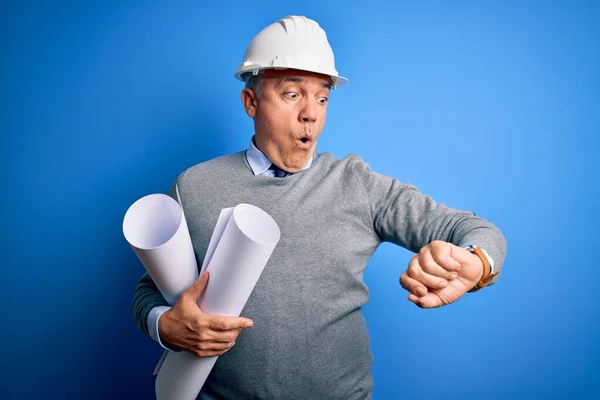 Middle age handsome grey-haired architect man wearing safety helmet holding blueprints Looking at the watch time worried, afraid of getting late