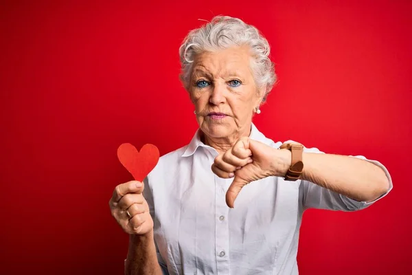 Senior beautiful woman holding paper heart standing over isolated red background with angry face, negative sign showing dislike with thumbs down, rejection concept