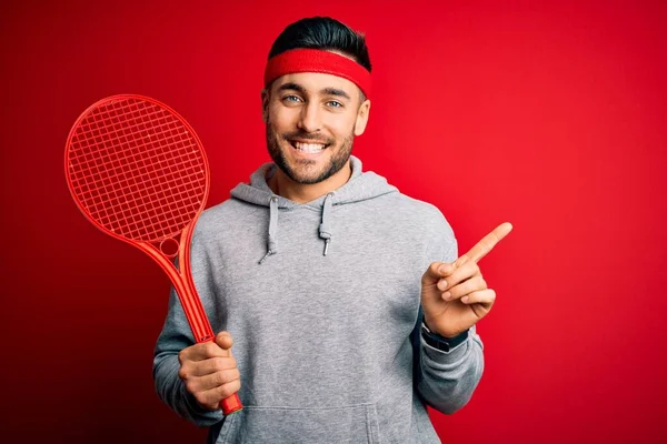 若いですハンサムなスポーツマン保持テニスラケット身に着けていますスポーツウェア以上赤背景非常に幸せなポインティングで手と指で側面 — ストック写真
