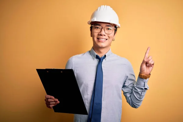 Młody Przystojny Chiński Inżynier Noszący Kask Bezpieczeństwa Trzymając Schowek Zaskoczony — Zdjęcie stockowe