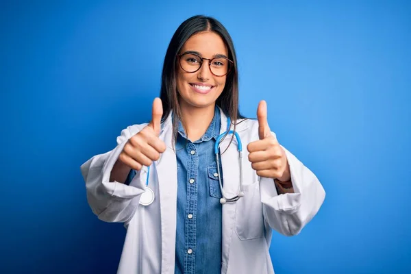青い背景の上に聴診器と眼鏡をかけている若い美しい医師の女性は 手で積極的なジェスチャーを行うことを承認し 親指を笑顔と成功のために幸せ 勝者のジェスチャー — ストック写真