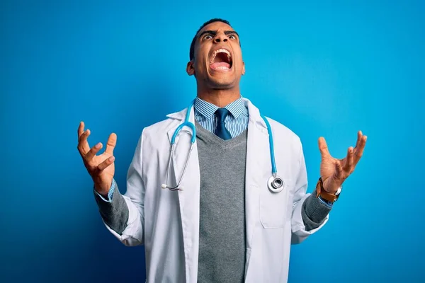 英俊的非洲裔美国医生 身穿外套和听诊器 身披蓝色背景 疯狂地喊叫着 怒气冲冲地举起双臂 沮丧的概念 — 图库照片