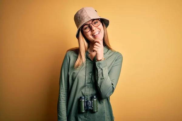 Beautiful blonde explorer woman with blue eyes wearing hat and glasses using binoculars looking confident at the camera with smile with crossed arms and hand raised on chin. Thinking positive.