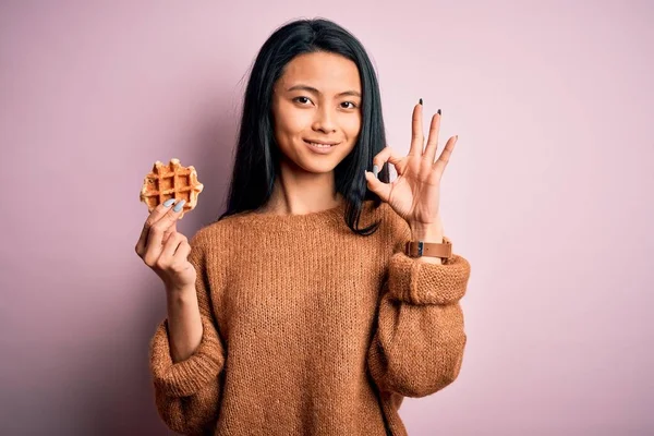 年轻美丽的中国女人拿着华夫饼站在孤零零的粉红背景上 手握手签 很好的象征 — 图库照片