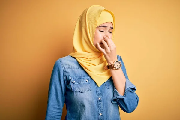 孤立した黄色の背景の上に立っているイスラム教徒のヒジャーブを身に着けている若い美しい少女は 鼻の上に指で息を保持し 臭いと嫌な 耐え難い匂いを嗅ぐ 匂いが悪い — ストック写真