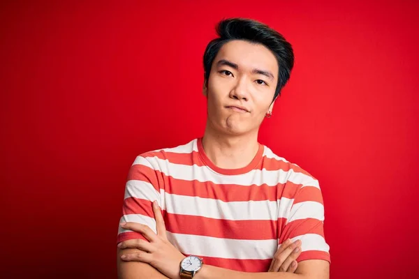 Young handsome chinese man wearing casual striped t-shirt standing over red background skeptic and nervous, disapproving expression on face with crossed arms. Negative person.