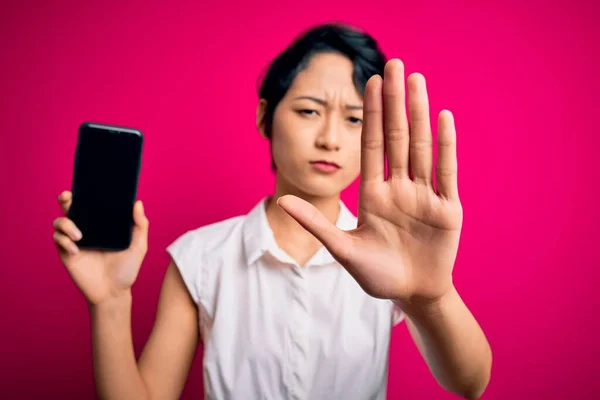 年轻美丽的亚洲女孩 手持智能手机 在孤立的粉色背景上展示屏幕 张开手做停止手势 表情严肃而自信 并做出防御姿态 — 图库照片