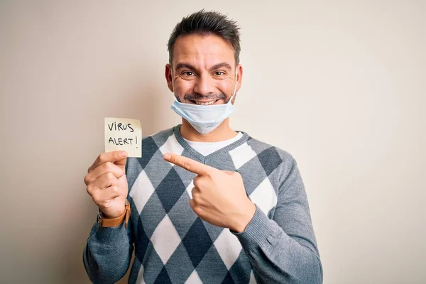 Young handsome man wearing medical mask holding paper with virus alert message very happy pointing with hand and finger