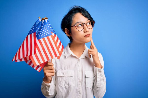 Young beautiful asian patriotic girl holding united states flags celebrating independence day serious face thinking about question, very confused idea