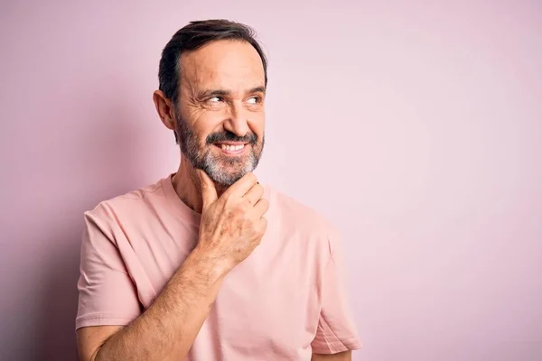 質問について考える顎の手で孤立したピンクの背景の上に立ってカジュアルなTシャツを着ている中年のホーリーマン 集中的な表現 思慮深い顔で微笑んだ 疑いの概念 — ストック写真