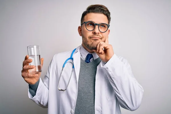 隔離された背景に新鮮な水のガラスを保持する医療用コートを身に着けている若い医者の男深刻な顔の質問について考えて 非常に混乱したアイデア — ストック写真