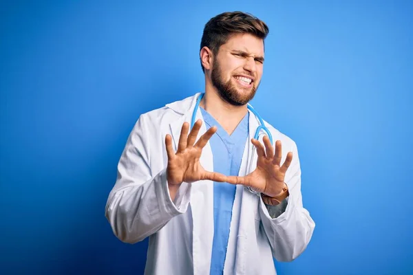 Young blond doctor man with beard and blue eyes wearing white coat and stethoscope disgusted expression, displeased and fearful doing disgust face because aversion reaction. With hands raised