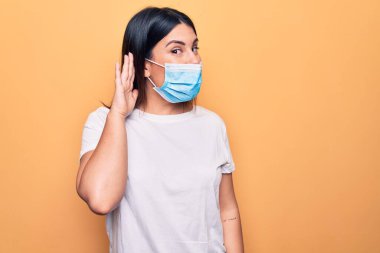Koronavirüs hastalığı için koruyucu maske takan genç bir kadın sarı arka planda sırıtarak dedikodu ya da dedikoduları dinliyor. Sağırlık kavramı.