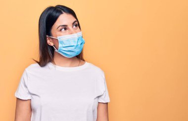 Koronavirüs hastalığı için koruyucu maske takan genç bir kadın sarı arka planda rahat bir görünüm ve kendine güvenen bir gülümseme takınıyor..