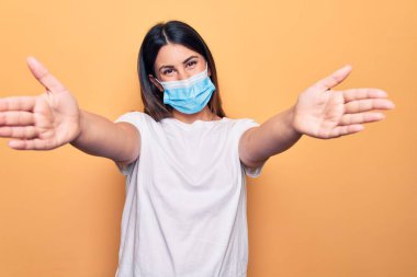 Koronavirüs hastalığı için koruyucu maske takan genç bir kadın sarı arka planda kameraya sarılmak için gülümseyerek bakıyor. Mutluluğu kucaklayan neşeli bir ifade..