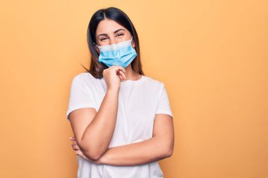 Koronavirüs hastalığı için koruyucu maske takan genç bir kadın sarı arka planda gülümsüyor ve kameraya kolları çapraz ve çenesi açık bakıyor. Olumlu düşünüyorum..