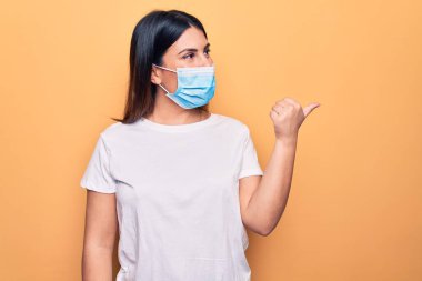 Koronavirüs hastalığı için koruyucu maske takan genç bir kadın sarı arka planda başparmağını yukarı kaldırıyor ve ağzı açık gülümsüyor.
