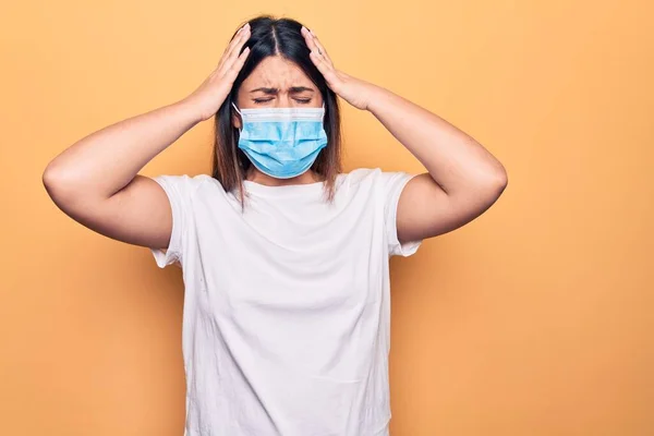 Young woman wearing protection mask for coronavirus disease over yellow background with hand on head, headache because stress. Suffering migraine.