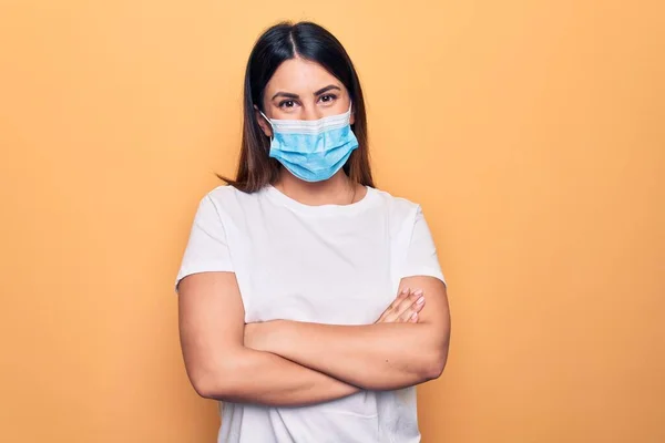 Young woman wearing protection mask for coronavirus disease over yellow background happy face smiling with crossed arms looking at the camera. Positive person.