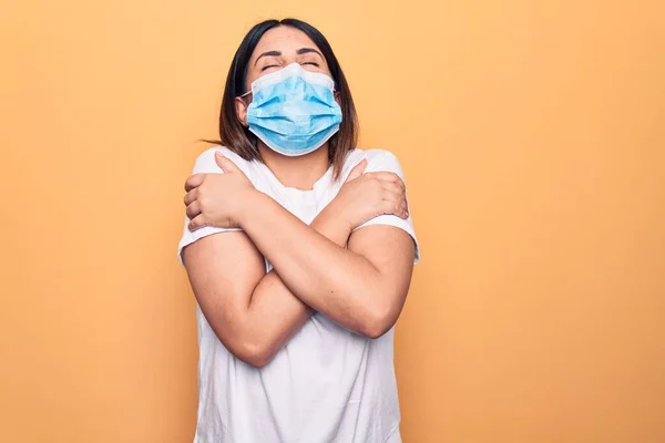 Young woman wearing protection mask for coronavirus disease over yellow background hugging oneself happy and positive, smiling confident. Self love and self care