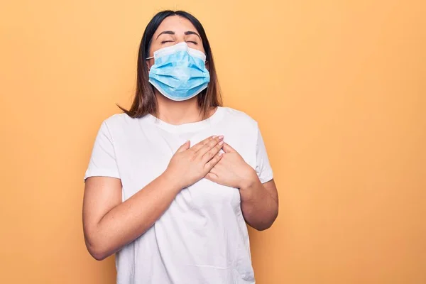 Young woman wearing protection mask for coronavirus disease over yellow background smiling with hands on chest, eyes closed with grateful gesture on face. Health concept.