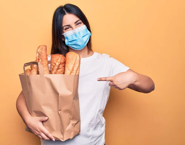 Young woman wearing protection mask for coronavirus holding take away paper bag with bread smiling happy pointing with hand and finger