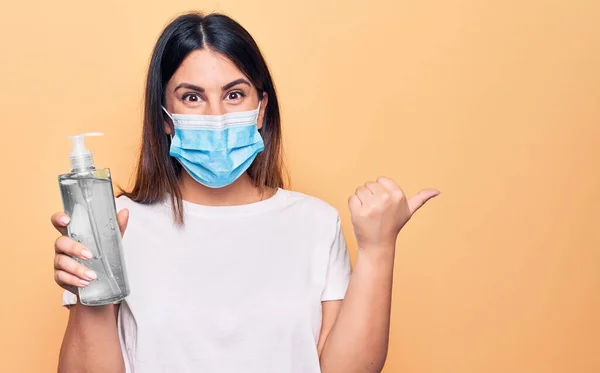 Young woman wearing protection mask for coronavirus holding hand sanitazer against virus disease pointing thumb up to the side smiling happy with open mouth