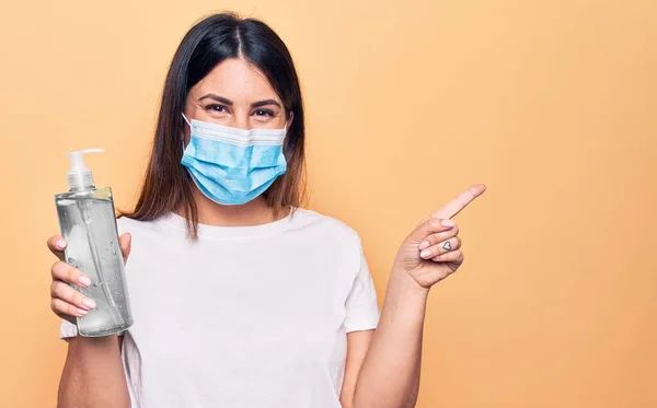 Young woman wearing protection mask for coronavirus holding hand sanitazer against virus disease smiling happy pointing with hand and finger to the side