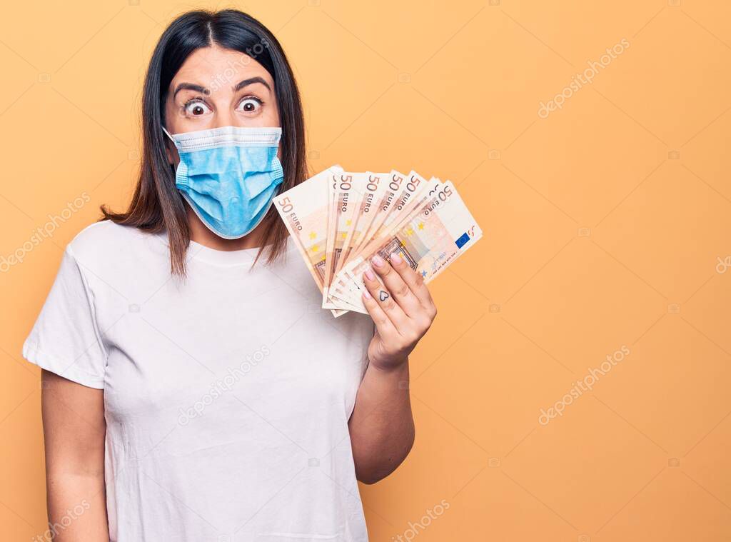 Young woman wearing protection mask for coronavirus holding 50 euros banknotes scared and amazed with open mouth for surprise, disbelief face