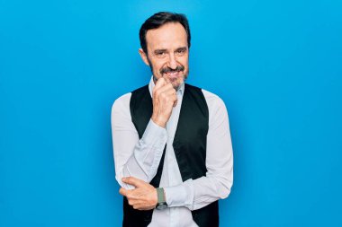 Soyutlanmış mavi arka plan üzerinde zarif gömlek ve yelek giyen yakışıklı bir adam kameraya kollarını kavuşturup çenesine dokunarak gülümsüyor. Olumlu düşünüyorum..