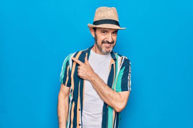 Orta yaşlı yakışıklı turist, tişört ve şapkayla mavi arka planda gülümsüyor.
