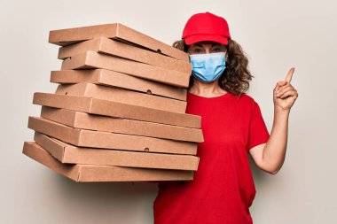 Orta yaşlı teslimatçı kadın, koronavirüs koruma maskesi takıyor ve elinde mukavva pizza kutuları tutarak mutlu bir şekilde gülümsüyor.
