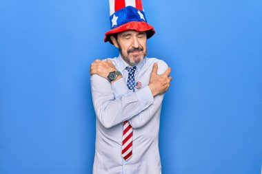 Orta yaşlı, vatansever, şapkalı ve kravatlı bir adam mavi arka planda mutlu, pozitif ve kendinden emin bir şekilde kendine sarılıyor. Sevgi ve özgüven.