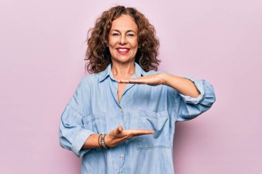 Orta yaşlı, güzel bir kadın pembe arka planda dikilmiş sıradan bir kot gömlek giyiyor. Ellerinde büyük ve büyük beden işareti, ölçü sembolü var. Kameraya bakıp gülümsüyor. Ölçüm konsepti.