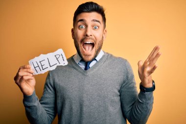 Genç aşırı çalışan iş adamı sarı arka planda kağıt tutarken mutlu ve heyecanlı, kazanan yüz ifadesi zaferi büyük bir gülümsemeyle kutluyor ve ellerini kaldırıyor.