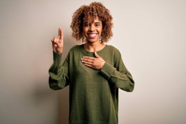 Güzel, kıvırcık saçlı, beyaz arka planda rahat süveter giyen, elleri göğsünde ve parmakları havada gülümseyen, sadakat yemini eden Afro-Amerikan bir kadın.