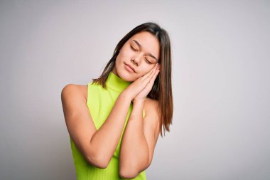 Genç, güzel esmer kız izole edilmiş beyaz arka planda günlük tişört giyip uyur, yorgun düşler kurar ve elleri ve gözleri kapalı gülümserken poz verir..